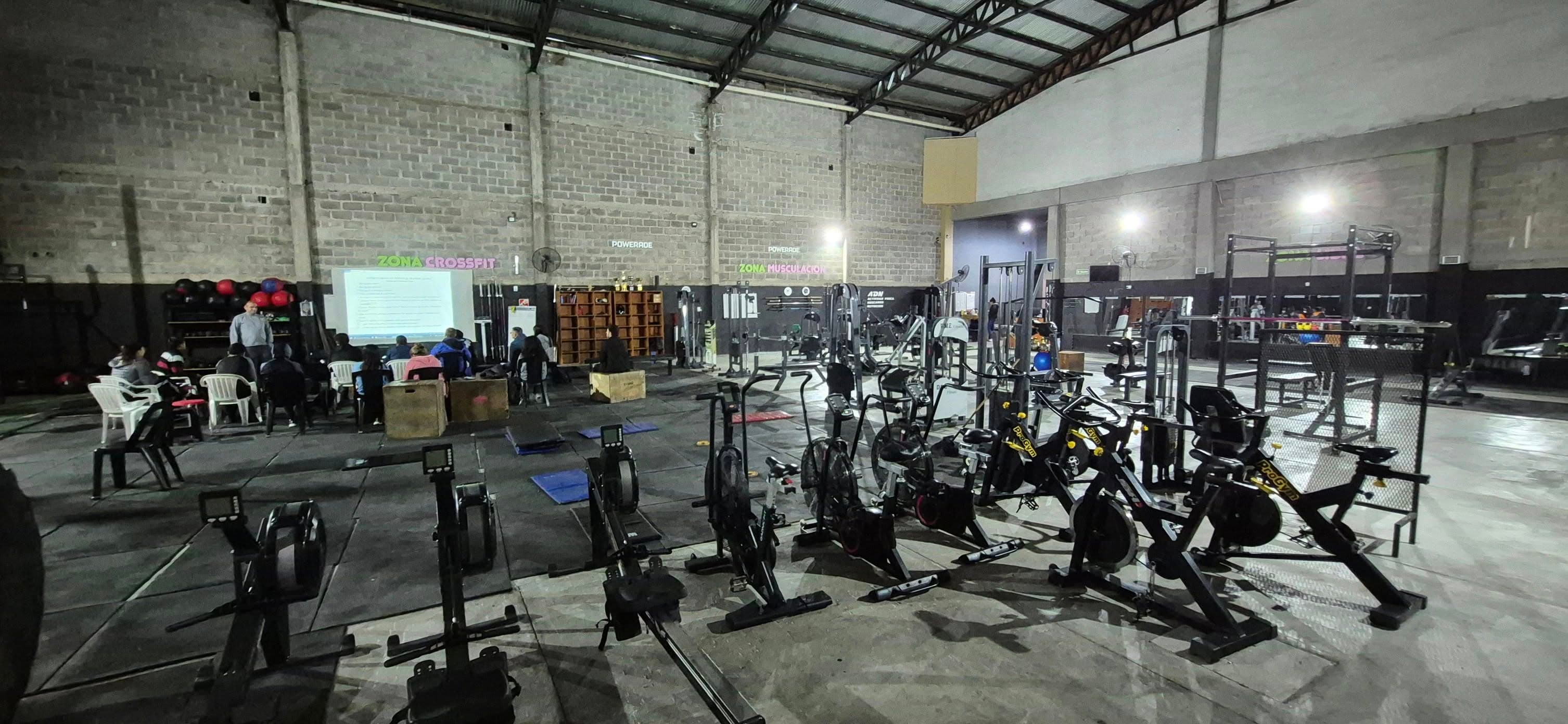 Gimnasio con máquinas de ejercicio y personas sentadas viendo una presentación en una pantalla.