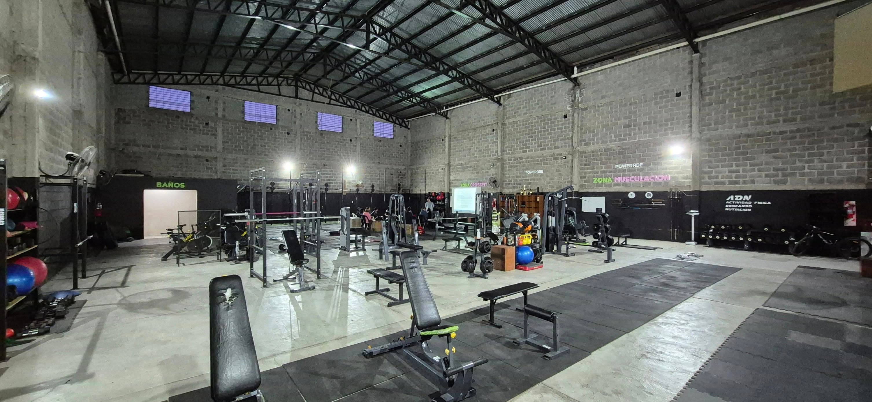 Amplio gimnasio industrial con bancos de pesas, estantes y equipos bajo un techo metálico alto.
